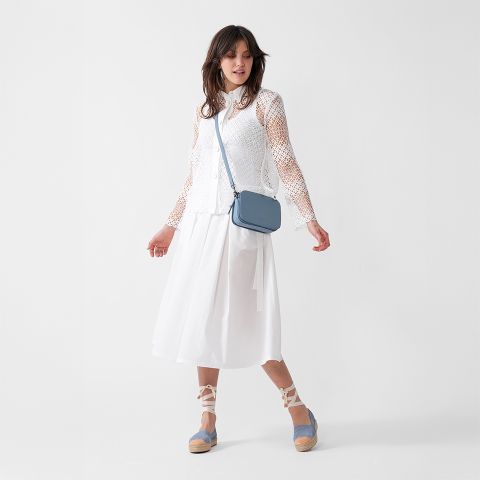 Eine Frau trägt ein langärmliges Oberteil aus weißer Spitze, einen weißen Midirock, blaue Espadrille-Wedges und eine hellblaue Umhängetasche und steht vor einem schlichten weißen Hintergrund.
