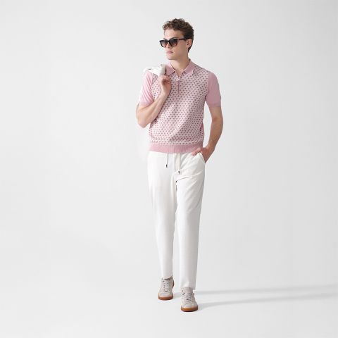 Ein Mann mit Sonnenbrille, einem hellrosa gemusterten Kurzarmhemd, weißen Hosen und grauen Turnschuhen steht vor einem schlichten weißen Hintergrund und hält eine weiße Jacke über der Schulter.