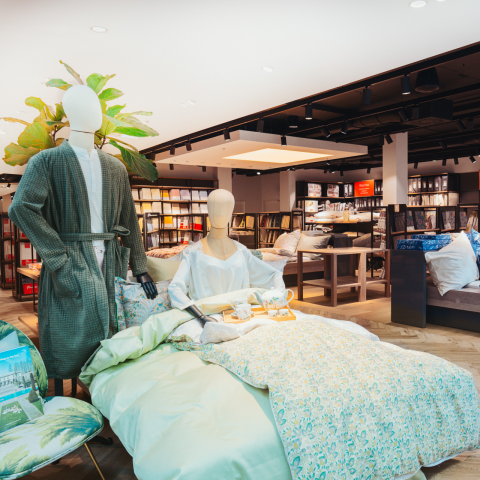 Ein Ladeninneres zeigt ein Bett mit geblümter Bettwäsche. Auf dem Bett stehen zwei Schaufensterpuppen, eine sitzt im Pyjama und die andere steht im Morgenmantel. In den Regalen daneben sind verschiedene Haushaltswaren ausgestellt und eine große Pflanze schmückt den Raum.