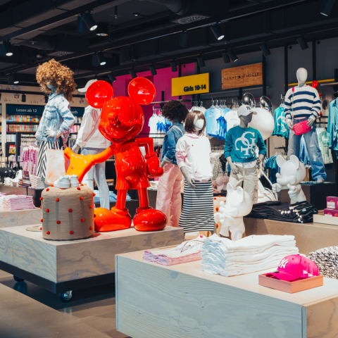Ein Schaufenster in einem Bekleidungsgeschäft mit Schaufensterpuppen in legerer Kleidung. Eine große, rote, cartoonartige Statue steht prominent zwischen ihnen. Das Geschäft ist hell erleuchtet und präsentiert verschiedene Kleidungsstücke auf Tischen und Regalen.
