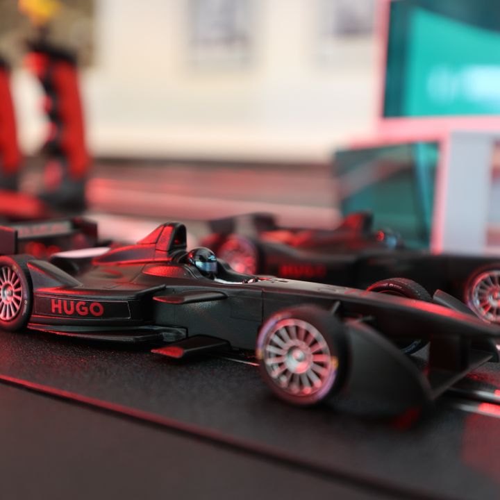 Nahaufnahme eines schwarzen Formel-1-Miniaturautos auf einer Rennstrecke mit dem Namen „Hugo“ an der Seite. Das Auto hat ein schnittiges Design mit sichtbaren Reifen und im Hintergrund ist ein weiteres Auto leicht verschwommen zu sehen.