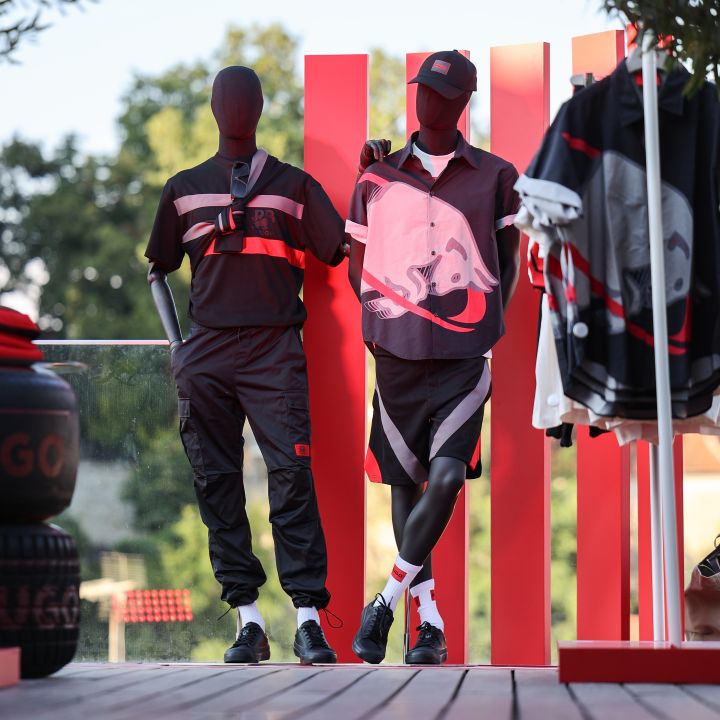 Zwei Schaufensterpuppen in sportlichen Outfits stehen auf einem Holzdeck vor einem rot-weiß gestreiften Hintergrund. Die linke Schaufensterpuppe trägt schwarz-rote Freizeitkleidung, während die rechte Schaufensterpuppe ein schwarz-rotes Sportensemble mit einer Kappe trägt. Im Hintergrund sind Bäume zu sehen.