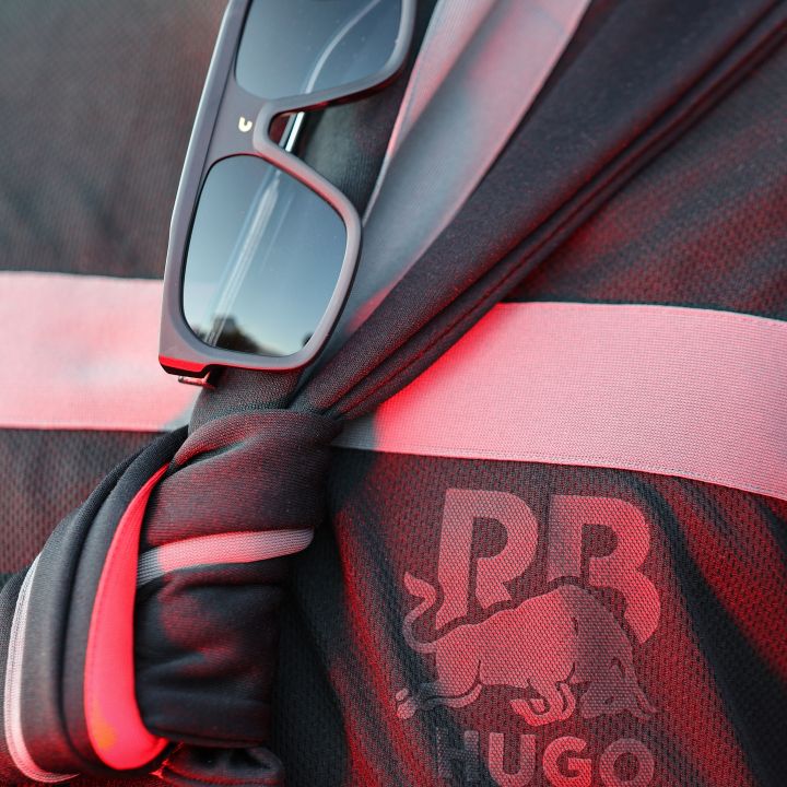Nahaufnahme eines schwarz-roten Sporttrikots mit den Logos „RB“ und „Hugo“ mit einem Stier. Im Kragen des Trikots, das mit einem geknoteten Stoffdetail versehen ist, steckt eine schwarze Sonnenbrille. Die Beleuchtung wirft einen warmen roten Farbton auf das Kleidungsstück.