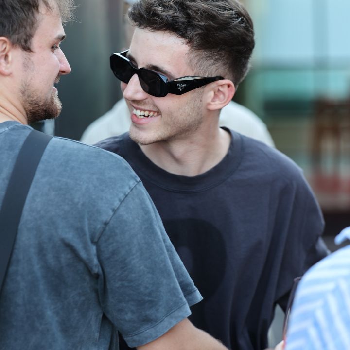 Zwei junge Männer lächeln sich im Freien an. Einer trägt eine schwarze Sonnenbrille und ein dunkles T-Shirt, während der andere einen hellen Bart hat und ein graues Hemd trägt. Sie scheinen sich zu begrüßen oder einen freundschaftlichen Moment miteinander zu verbringen.