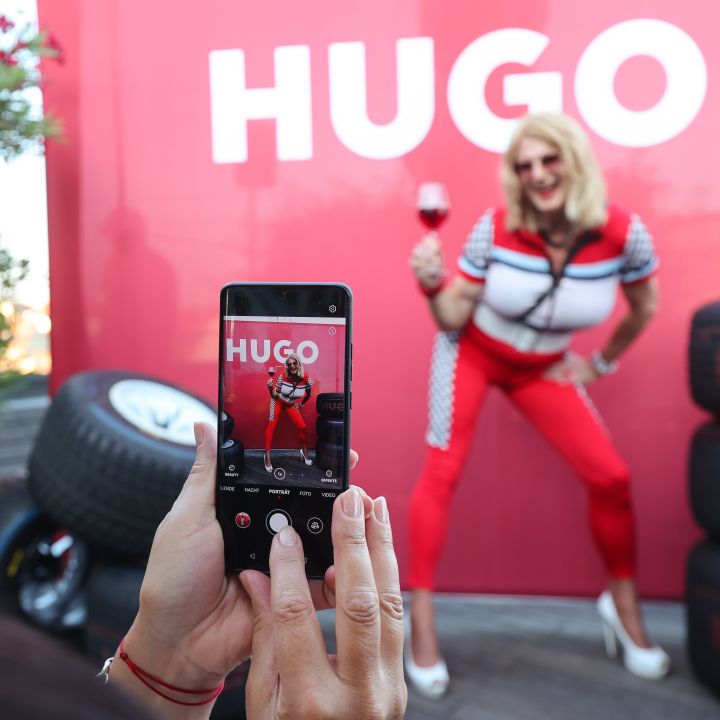 Person in rot-weißem Outfit posiert mit einem Weinglas vor einem roten Hintergrund mit dem Text „HUGO“. Eine andere Person macht ein Foto mit einem Telefon. Auf der rechten Seite sind Reifen gestapelt.