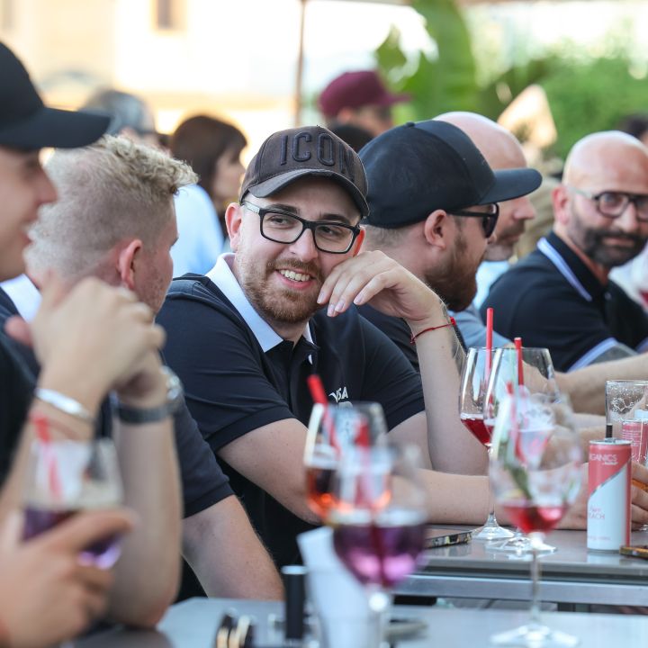 Eine Gruppe von Menschen sitzt an einem langen Tisch im Freien und genießt Getränke in Gläsern mit roter Flüssigkeit. Sie sind leger gekleidet, einige tragen Mützen und Sonnenbrillen und scheinen in ein Gespräch vertieft zu sein und zu lächeln.