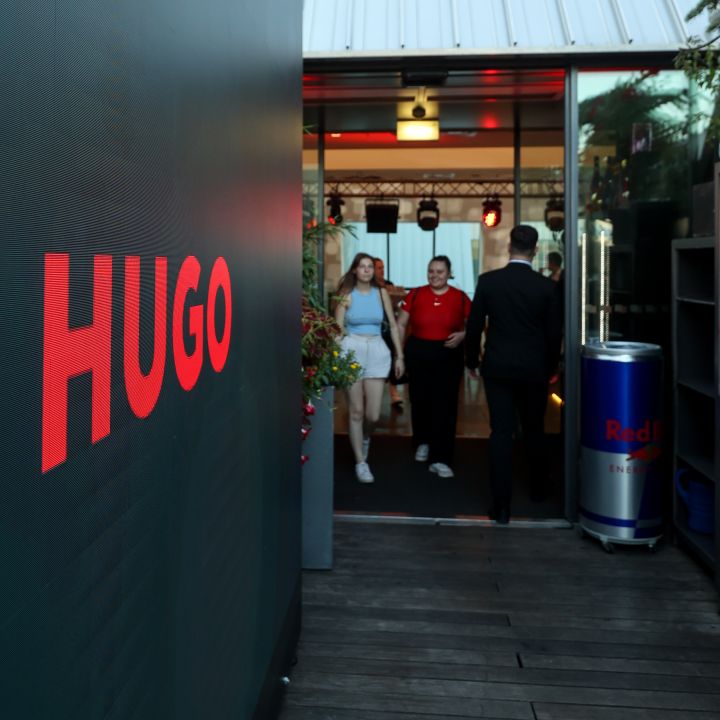 Eingang zu einem Veranstaltungsort mit einem großen „HUGO“-Schild an einer Wand. Drei Personen gehen hinein: eine Frau in einem weißen Outfit, ein Mann in einem roten Hemd und ein Mann in einem dunklen Anzug. Rechts ist eine Red Bull-Kühlbox und darüber ein schräges Dach.