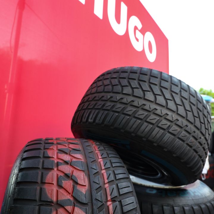 Drei gestapelte Autoreifen mit sichtbaren Profilspuren stehen vor einer roten Wand, auf der in großen weißen Buchstaben das Wort „HUGO“ steht.