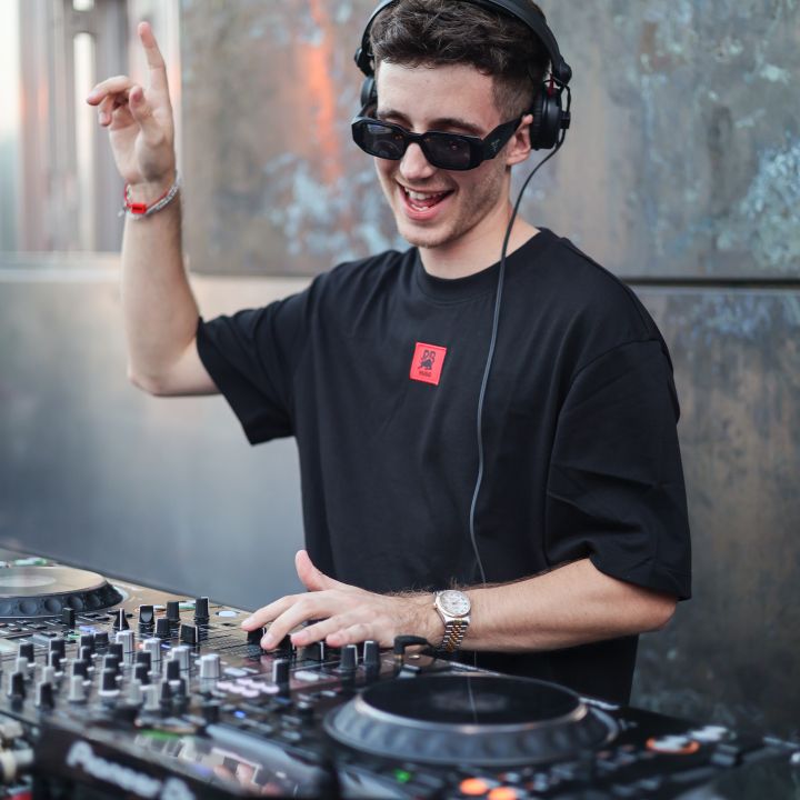 Ein DJ mit Sonnenbrille und Kopfhörern spielt voller Energie Musik auf einem Mischpult. Er hebt eine Hand und lächelt. Er trägt ein schwarzes T-Shirt mit rotem Muster. Im Hintergrund ist eine strukturierte Metallwand zu sehen.