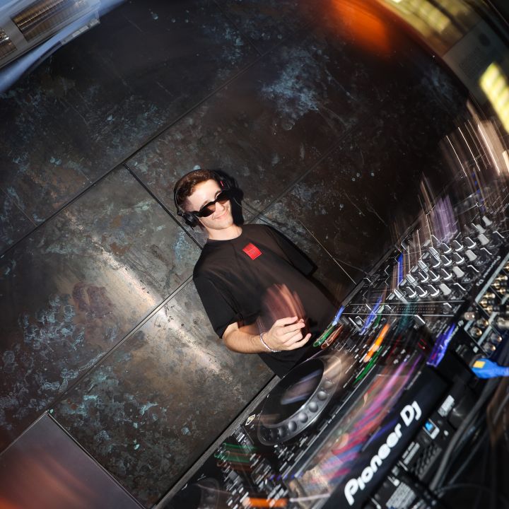 Ein DJ mit Sonnenbrille und Kopfhörern legt an einem Mischpult auf. Die Szene wird von warmem Licht erhellt und es gibt verschwommene Bewegungseffekte, die dem Bild dynamische Energie verleihen.