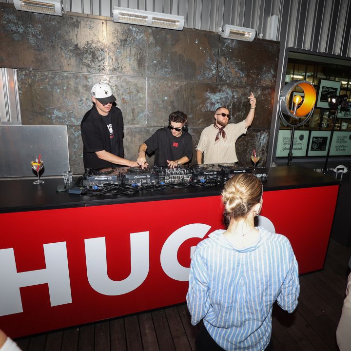DJ tritt bei einer Veranstaltung im Freien hinter einem roten Tresen mit der Aufschrift „HUGO“ auf. Drei Personen stehen am DJ-Pult, Zuschauer sind versammelt. Die Kulisse ist mit industriellen Elementen und warmer Beleuchtung gestaltet.