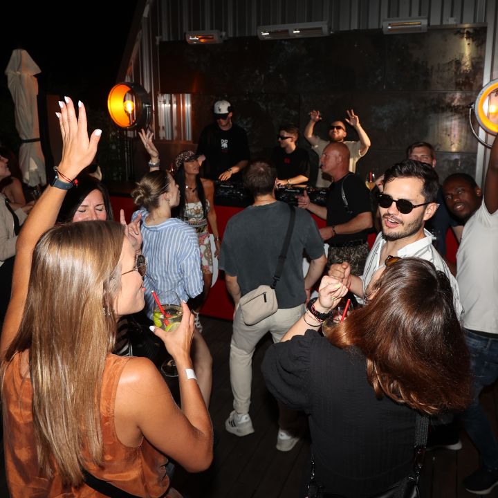 Eine lebhafte Partyszene mit Menschen, die tanzen und Musik genießen. Mehrere Personen haben ihre Hände erhoben und im Hintergrund läuft ein DJ. Neonlichter und eine Mischung aus lässiger und eleganter Kleidung tragen zur lebhaften Atmosphäre bei.