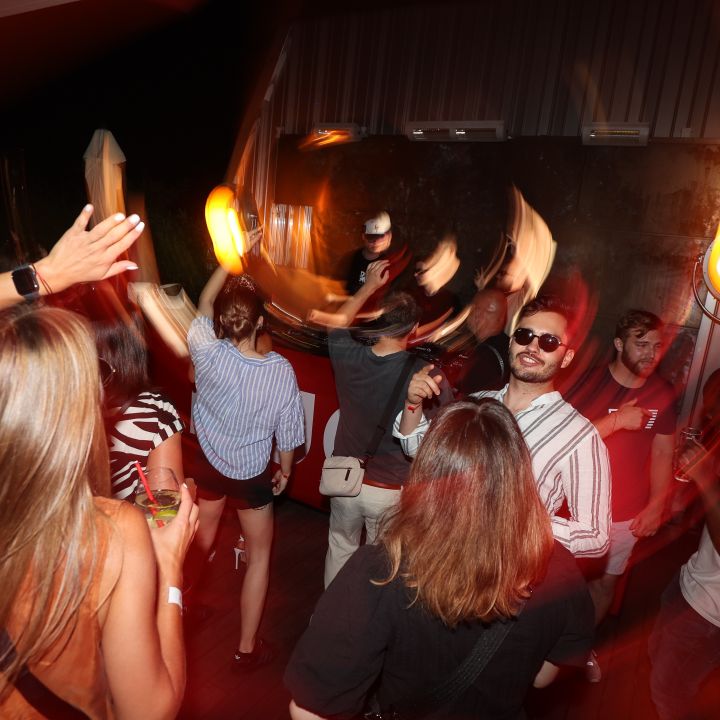 Menschen tanzen energisch in einem schwach beleuchteten Club mit warmer Beleuchtung. Bewegungsunschärfe fängt die lebhafte Atmosphäre ein. Eine Person im Vordergrund hat einen Arm erhoben, während andere sich unterhalten und die Musik genießen.