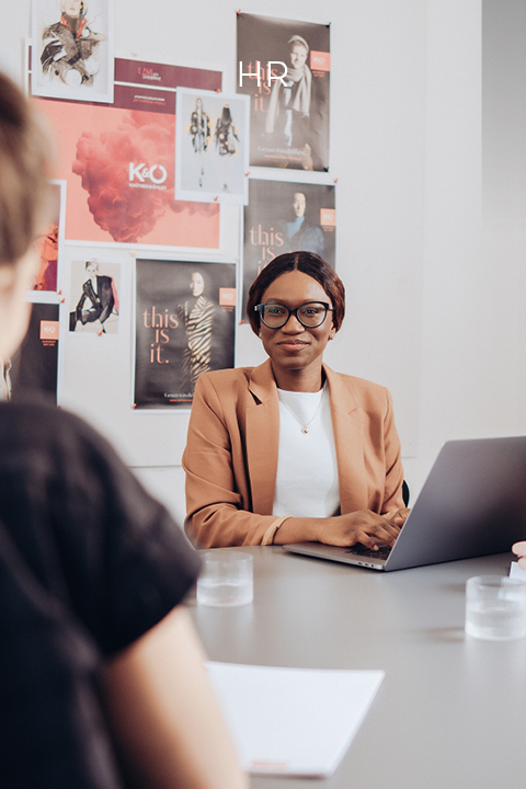 Eine Person in einem hellbraunen Blazer und mit Brille sitzt mit einem Laptop an einem Tisch und nimmt an einer Besprechung teil. Im Vordergrund sind verschwommene Figuren zu sehen. Im Hintergrund hängen Poster an der Wand.