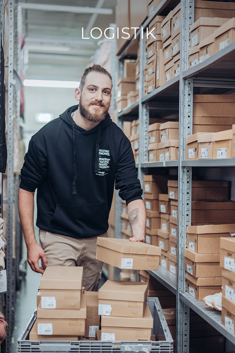 Ein Mann mit Bart und schwarzem Kapuzenpullover ordnet Kartons auf Metallregalen in einem Lagerhaus. Über ihm steht das Wort „LOGISTIKI“. Die Regale sind mit ähnlichen Kartons gefüllt.