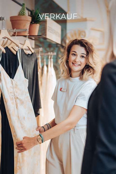 Eine Verkäuferin mit schulterlangem blondem Haar lächelt und hält ein geblümtes Kleid in einem Bekleidungsgeschäft. Darüber hängt ein Schild mit der Aufschrift „VERKAUF“. Das Geschäft hat warmes Licht und Topfpflanzen.