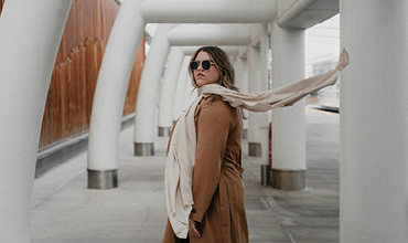 Eine Person mit Sonnenbrille, braunem Mantel und wallendem Schal steht in einem leeren Außenkorridor mit großen weißen Säulen und Holzvertäfelung im Hintergrund.