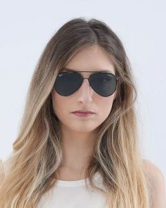 Frau mit langen, welligen blonden Haaren, dunkler Pilotensonnenbrille und weißem Oberteil. Sie hat einen neutralen Gesichtsausdruck und blickt vor einem schlichten hellen Hintergrund in die Kamera.