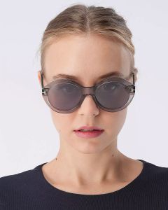 Eine Person mit einer runden schwarzen Sonnenbrille und einem schwarzen Hemd blickt vor einem schlichten weißen Hintergrund nach vorne. Die Haare sind zurückgekämmt.