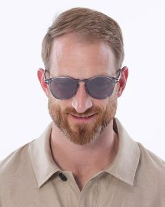 Ein Mann mit kurzen braunen Haaren und Bart, einer Sonnenbrille und einem beigen Hemd mit Kragen lächelt vor einem schlichten weißen Hintergrund.