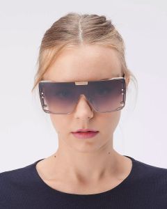 Eine Person mit zurückgebundenem hellbraunem Haar trägt eine übergroße, rechteckige Sonnenbrille mit dickem Rahmen. Sie trägt ein dunkles, schlichtes Oberteil und steht vor einem schlichten weißen Hintergrund.