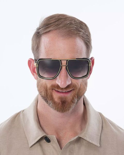 Ein Mann mit kurzen Haaren und Bart trägt eine große, quadratische Sonnenbrille mit dunklen Gläsern. Er trägt ein beiges Hemd mit Kragen und lächelt. Der Hintergrund ist schlicht weiß.