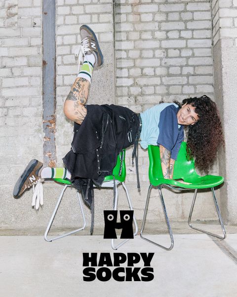 Eine Person mit lockigem Haar und Tattoos posiert verspielt auf zwei grünen Stühlen. Sie trägt ein blaues Hemd, einen schwarzen Rock, gestreifte Socken und Turnschuhe. Darunter sind in fetten Buchstaben die Worte „Happy Socks“ zu sehen. Im Hintergrund ist eine Backsteinmauer zu sehen.