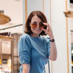 Eine Frau mit braunem Haar ist drinnen, lächelt und hebt eine braune Sonnenbrille. Sie trägt einen blauen Pullover und eine Uhr und steht vor einem Spiegel mit verschwommenen Hintergrundelementen.