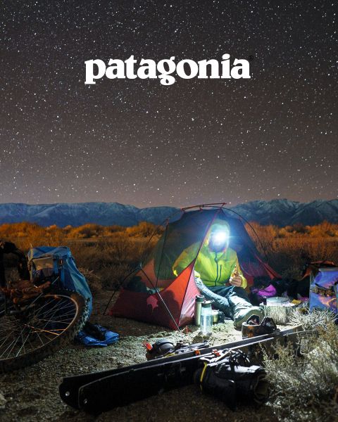 Eine Person mit Stirnlampe sitzt in einem kleinen beleuchteten Zelt unter einem Sternenhimmel an einem Lagerfeuer. In der Nähe sind Fahrräder, Outdoor-Ausrüstung und oben ein Patagonia-Logo zu sehen. Im Hintergrund sind kahle Landschaft und eine Bergkette zu sehen.