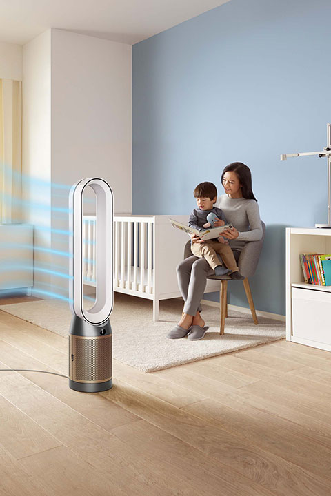 Eine Frau sitzt auf einem Stuhl und liest einem Kind in einem Kinderzimmer mit Kinderbett, Bücherregal und Lampe vor. Im Vordergrund stößt ein Luftreiniger in schlankem, modernem Design Luftströme aus. Der Raum hat helle Holzböden und sanfte blaue Wände.