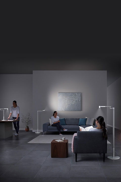 In einem schwach beleuchteten Wohnzimmer sind drei Personen mit unterschiedlichen Tätigkeiten beschäftigt: eine steht an einem Schreibtisch, eine andere sitzt mit einem Buch auf einem Sofa und eine dritte sitzt lesend auf einem Stuhl. Modernes Dekor und Stehlampen erhellen den Raum.