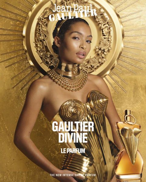 Ein Model in einem goldenen Korsettkleid posiert vor einem goldenen dekorativen Hintergrund und hält eine Parfümflasche. Der Text lautet „Jean Paul Gaultier“, „Gaultier Divine“ und „Le Parfum The New Intense Eau de Parfum“.