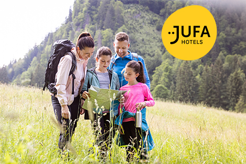 Eine vierköpfige Familie mit Rucksäcken steht auf einer Wiese und schaut auf eine Karte. Sie sind von Bäumen und Hügeln umgeben. Das Logo der JUFA Hotels befindet sich in der oberen rechten Ecke. Der Himmel ist klar und sonnig.