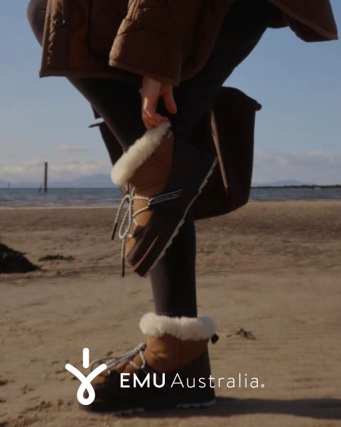 Eine Person in braunen, pelzgefütterten EMU Australia-Stiefeln und einem braunen Mantel steht an einem Sandstrand und hält einen Fuß hinter sich hoch. Das EMU Australia-Logo erscheint am unteren Rand des Bildes.