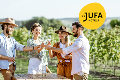 Vier Personen stoßen an einem sonnigen Tag im Freien neben einem Weinberg mit Weingläsern an. Sie sind leger gekleidet und lächeln. In der oberen rechten Ecke befindet sich ein gelbes „JUFA Hotels“-Logo. Vor ihnen steht ein Holztisch mit einer Weinflasche.
