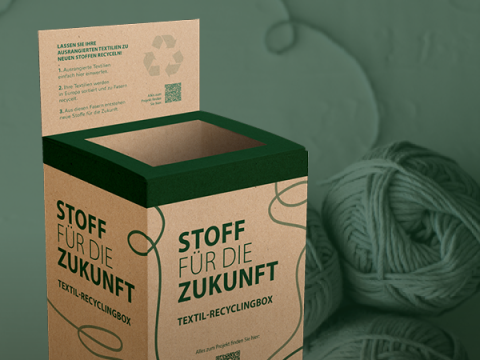 Eine Recyclingbox aus Pappe mit der Aufschrift „Stoff für die Zukunft“ und einem Recyclingsymbol. Auf der Oberseite ist eine Anleitung aufgedruckt. Daneben liegt auf einer grünen Fläche ein Knäuel grünes Garn.