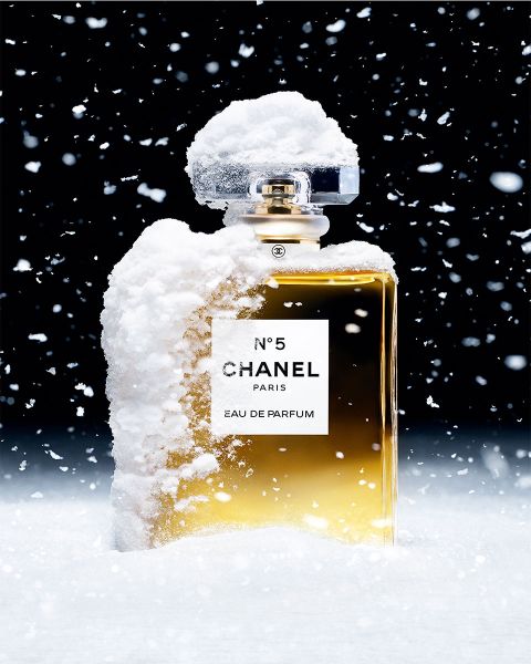 Ein mit Schnee bestäubter Chanel No. 5-Parfümflakon steht aufrecht vor einem dunklen Hintergrund. Schneeflocken fallen sanft um den Flakon herum und erzeugen eine winterliche Szene. Die goldene Flüssigkeit im transparenten Flakon kontrastiert mit dem weißen Schnee.