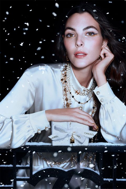 Eine Frau in einer seidigen weißen Bluse, geschmückt mit mehreren Perlen- und Goldketten, blickt nach vorne, während um sie herum Schnee fällt. Sie hält eine kleine Chanel-Geschenkschachtel in der Hand. Im Hintergrund ist eine dunkle, verschneite Nacht zu sehen.
