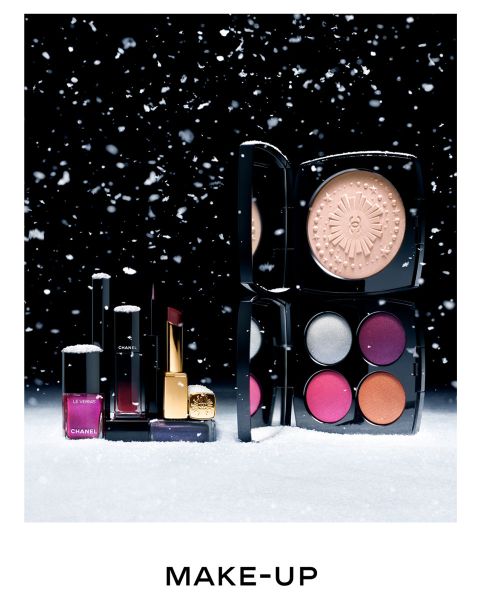 Eine Sammlung von Make-up-Produkten auf einer schneebedeckten Oberfläche mit schwarzem Hintergrund. Enthält ein Kompaktset mit vier bunten Lidschatten, Textmarker, Lippenstiften und Nagellack. Schneeflocken sind in der Luft zu sehen. Darunter steht das Wort „MAKE-UP“.