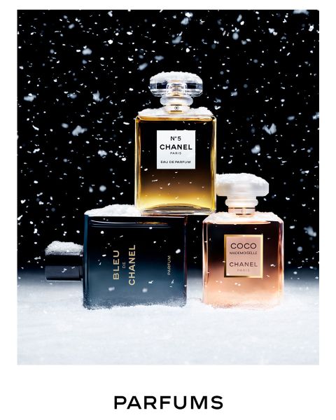 Drei Chanel-Parfümflaschen, mit Schnee bedeckt, sind vor einem schwarzen Hintergrund mit fallenden Schneeflocken angeordnet. Die Flaschen tragen die Aufschriften N°5, BLEU und COCO MADEMOISELLE. Darunter ist das Wort „PARFUMS“ zu sehen.