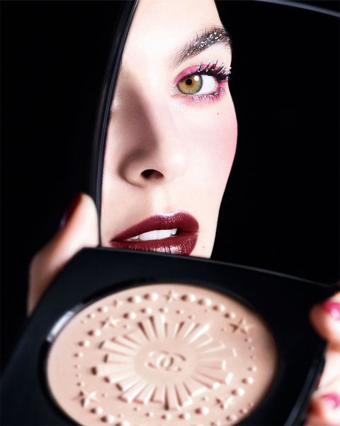 Eine Frau mit auffälligem Make-up hält einen Kompaktspiegel nah an ihr Gesicht. Sie trägt dunkelroten Lippenstift, schimmernden Lidschatten und betonte Augenbrauen. Der Kompaktspiegel enthält ein Kompaktpuder mit strukturierter Oberfläche. Der Hintergrund ist schwarz.