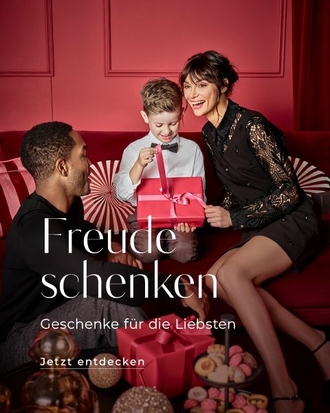 Eine Familie mit einem Mann, einer Frau und einem Kind sitzt in einem in Rot gehaltenen Raum. Das Kind öffnet aufgeregt eine rote Geschenkbox. Der Text auf dem Bild lautet: „Freude schenken, Geschenke für die Liebsten, Jetzt entdecken.“.