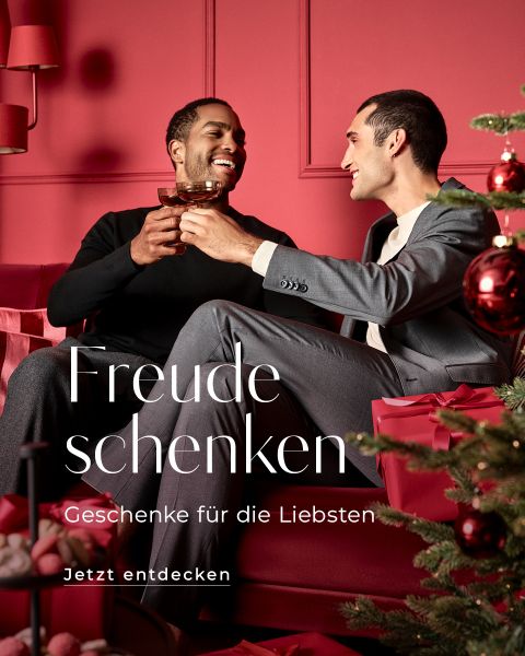 Zwei Männer sitzen auf einer roten Couch in einem festlich geschmückten Raum, stoßen mit Gläsern an und lächeln. Sie sind von Weihnachtsschmuck und einem Baum umgeben. Der deutsche Text auf dem Bild lautet: „Freude schenken. Geschenke für die Liebsten. Jetzt entdecken.“