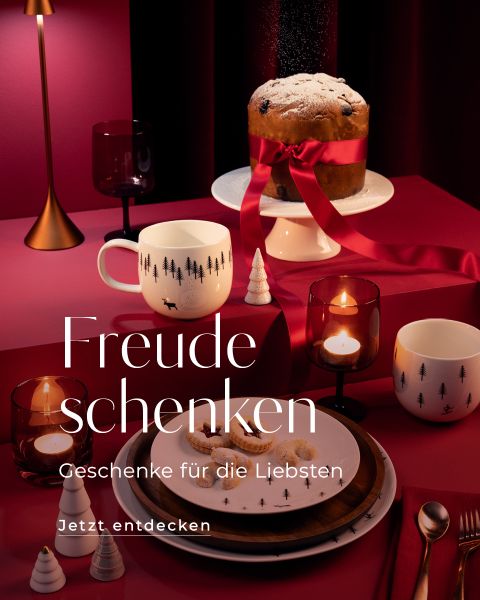 Ein gemütlich gedeckter Feiertagstisch mit brennenden Kerzen, Tassen und Tellern mit Baummotiven. Ein mit einer roten Schleife umwickelter und mit Zucker bestäubter Panettone steht auf einem Etagerenständer. Die Textüberlagerungen lauten „Freude schenken“ und „Geschenke für die Liebsten“.