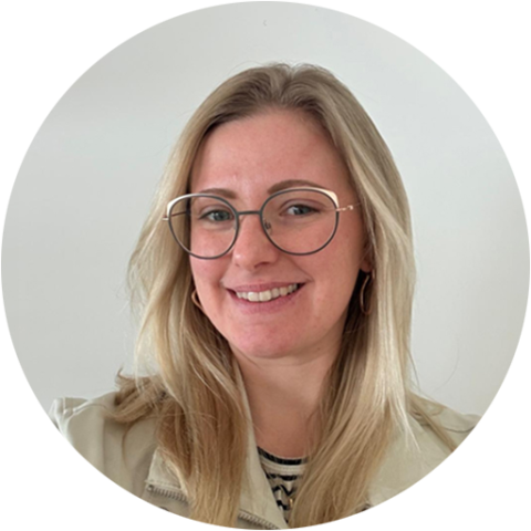 Eine Frau mit langen blonden Haaren, die eine Brille und eine helle Jacke trägt, lächelt vor einem schlichten weißen Hintergrund.