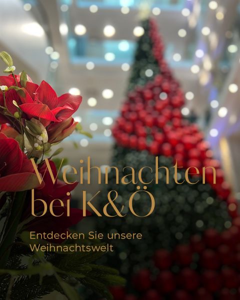 Ein hoher, mit roten Ornamenten geschmückter Weihnachtsbaum in einem Einkaufszentrum. Rote Blumen stehen im Vordergrund. Der Text lautet „Weihnachten bei K&O“ und „Entdecken Sie unsere Weihnachtswelt“ und suggeriert eine festliche Feiertagsstimmung.