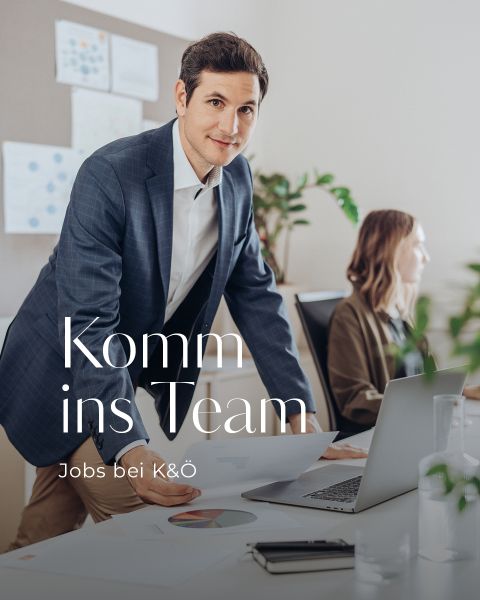 Ein Mann im Anzug lehnt an einem Schreibtisch in einem Büro und hält Papiere in der Hand. Eine Frau sitzt am Schreibtisch vor einem Laptop. Der Text auf dem Bild lautet „Komm ins Team – Jobs bei K&O“. Im Hintergrund sind Diagramme und Pflanzen zu sehen.