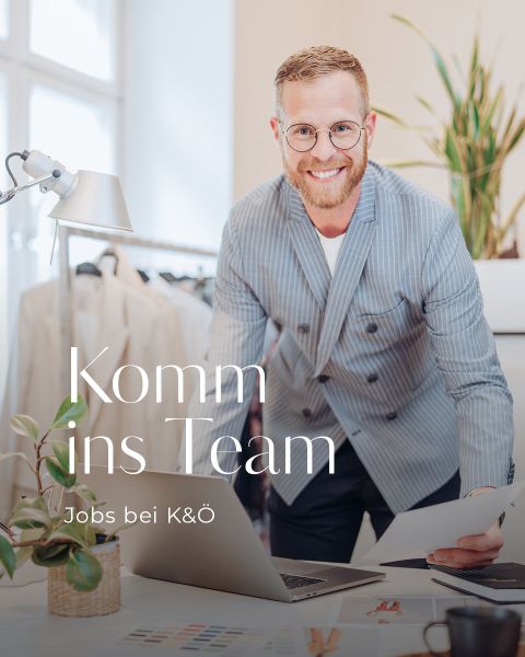 Ein Mann mit Brille und Bart, der einen gestreiften Blazer trägt, beugt sich nach vorne über einen Schreibtisch mit Papieren und einer Pflanze. Auf dem Bild steht „Komm ins Team“ und „Jobs bei K&Ö“. Im Hintergrund helles Büroambiente.