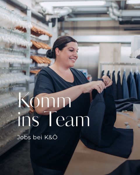 Eine lächelnde Person sortiert Kleidung in einem Arbeitsbereich mit Kleiderständern im Hintergrund. Der Text auf dem Bild lautet: „Komm ins Team, Jobs bei K&O.“