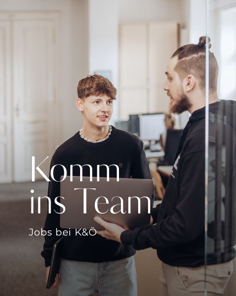 Zwei Männer stehen in einem Büro und unterhalten sich. Einer hält einen Laptop in der Hand. Auf dem Bild steht: „Komm ins Team – Jobs bei K&O“. Im Hintergrund sind Schreibtische und Computer zu sehen.
